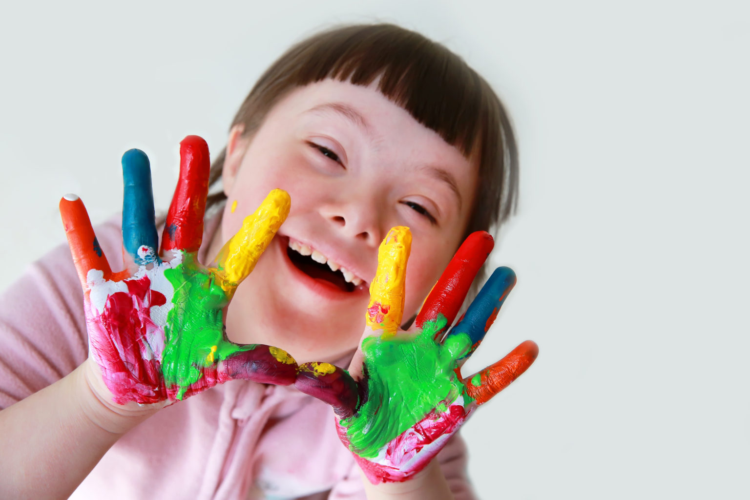 Image of a smiling litle girl with Down Syndrome. She is holding up her hands and they are covered with an assortemnt of colorful paitnts